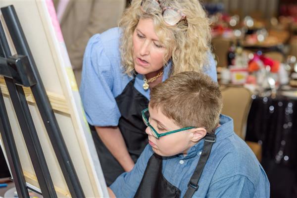 A student and an artist are focused on painting a masterpiece. The student wears glasses and an apron while the artist crouches down beside him. 
